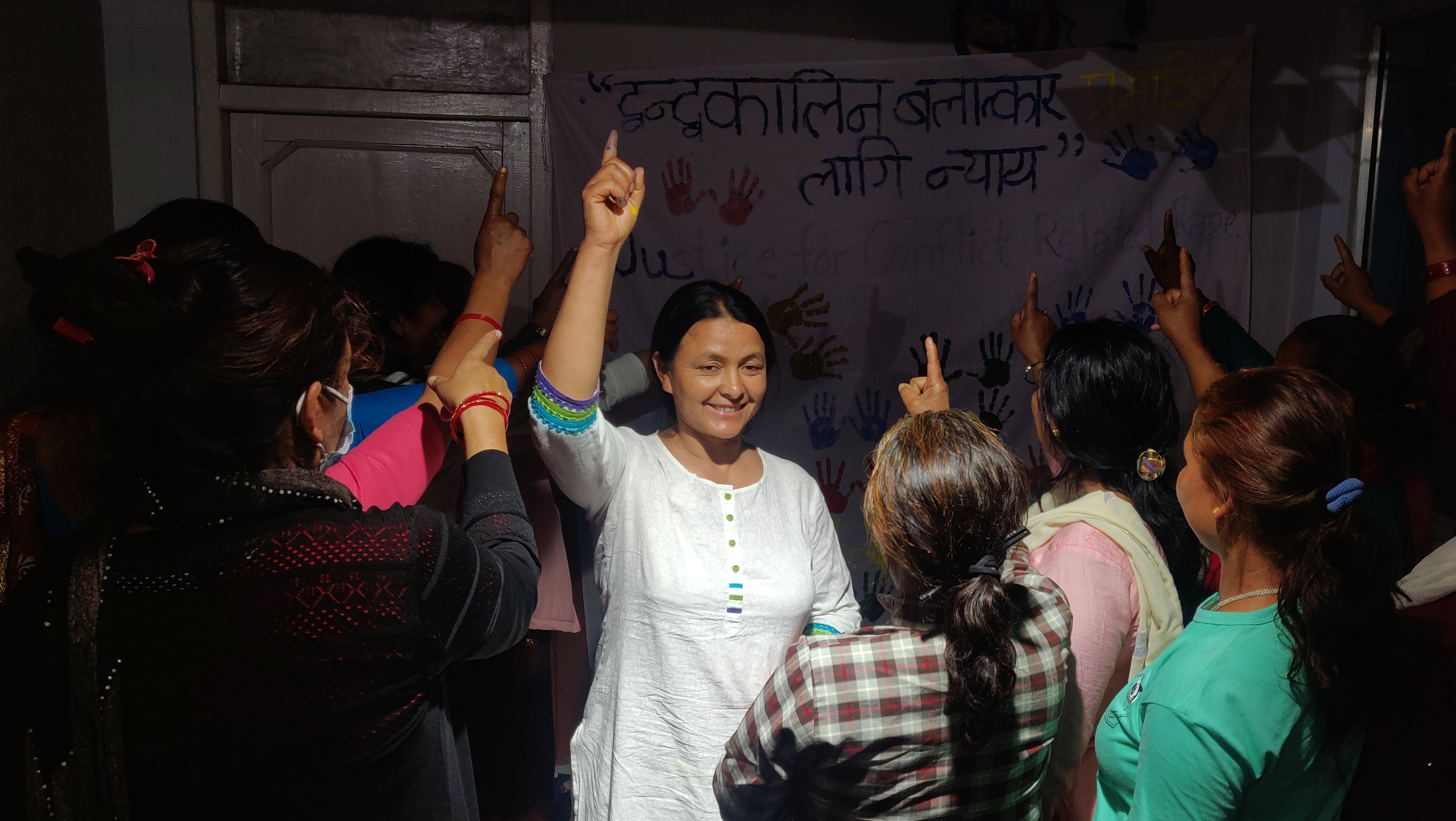 A group of women gathers, raising their index fingers in unison. At the center, a smiling Devi Khadka leads the gesture. Behind them, a banner with Nepali text and colorful handprints is visible.