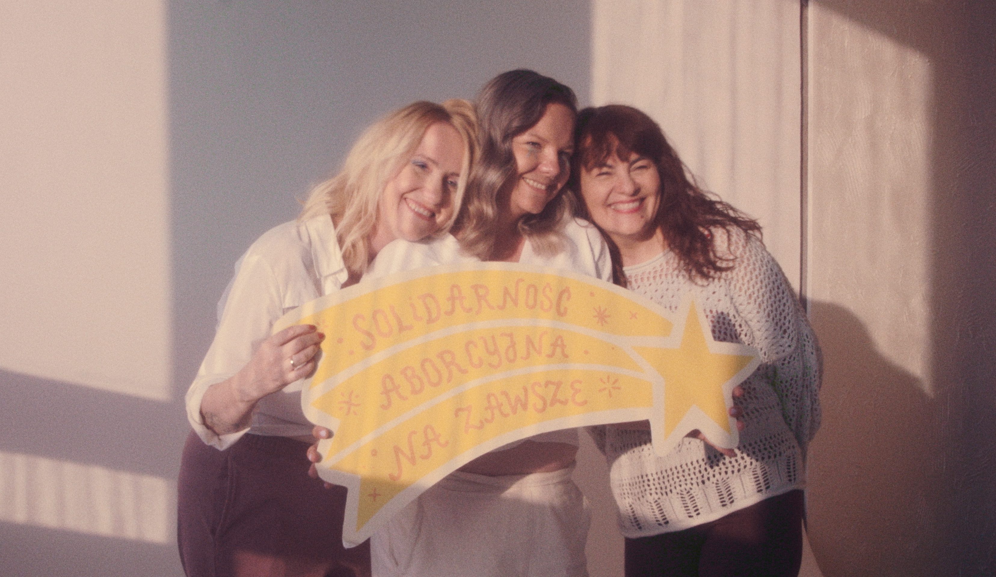 Three smiling women stand close together, holding a yellow sign shaped like a shooting star. The sign displays the Polish phrase 