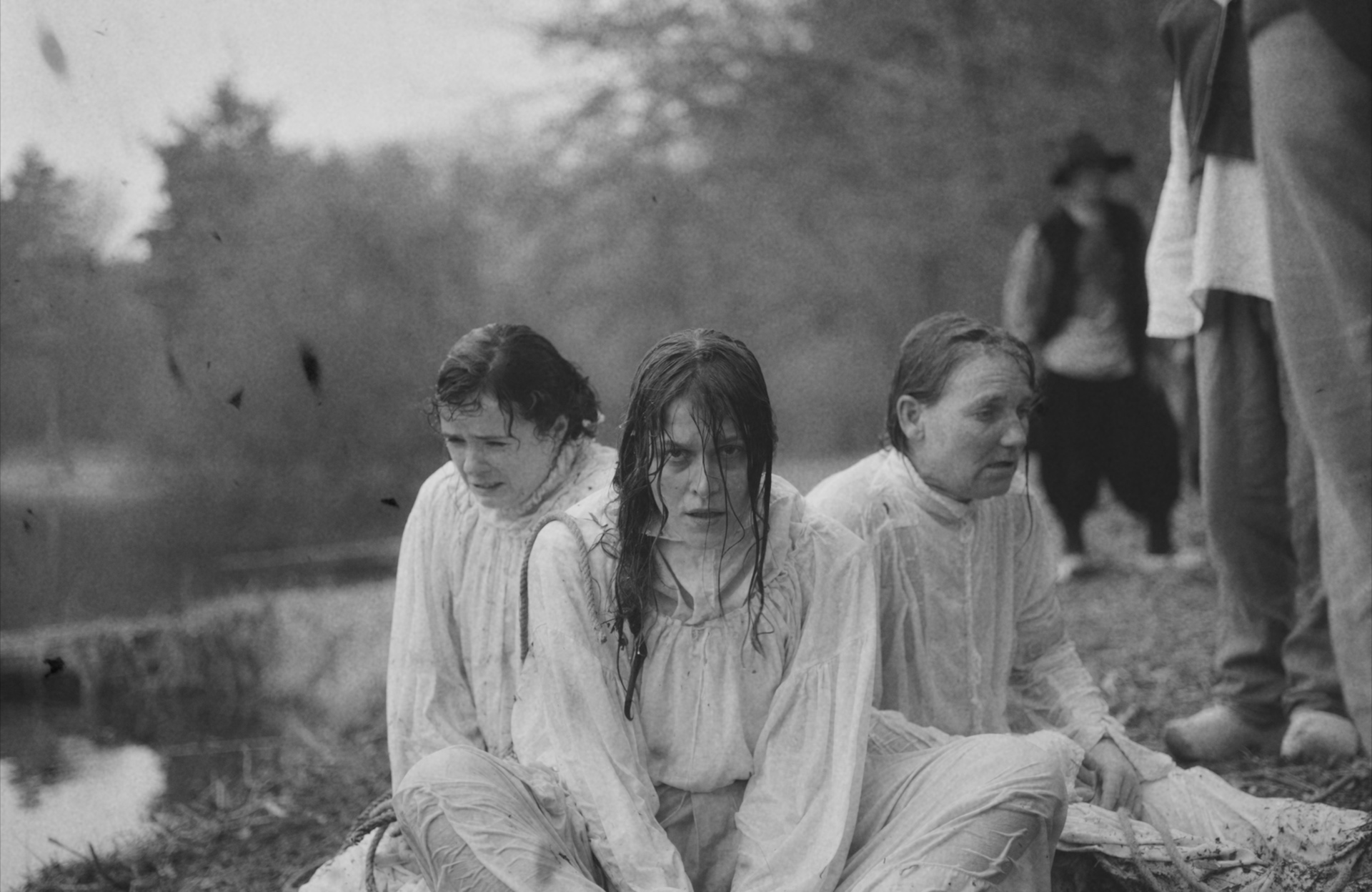 Three women in wet, white clothing sit near a body of water, surrounded by trees and onlookers in historical attire.