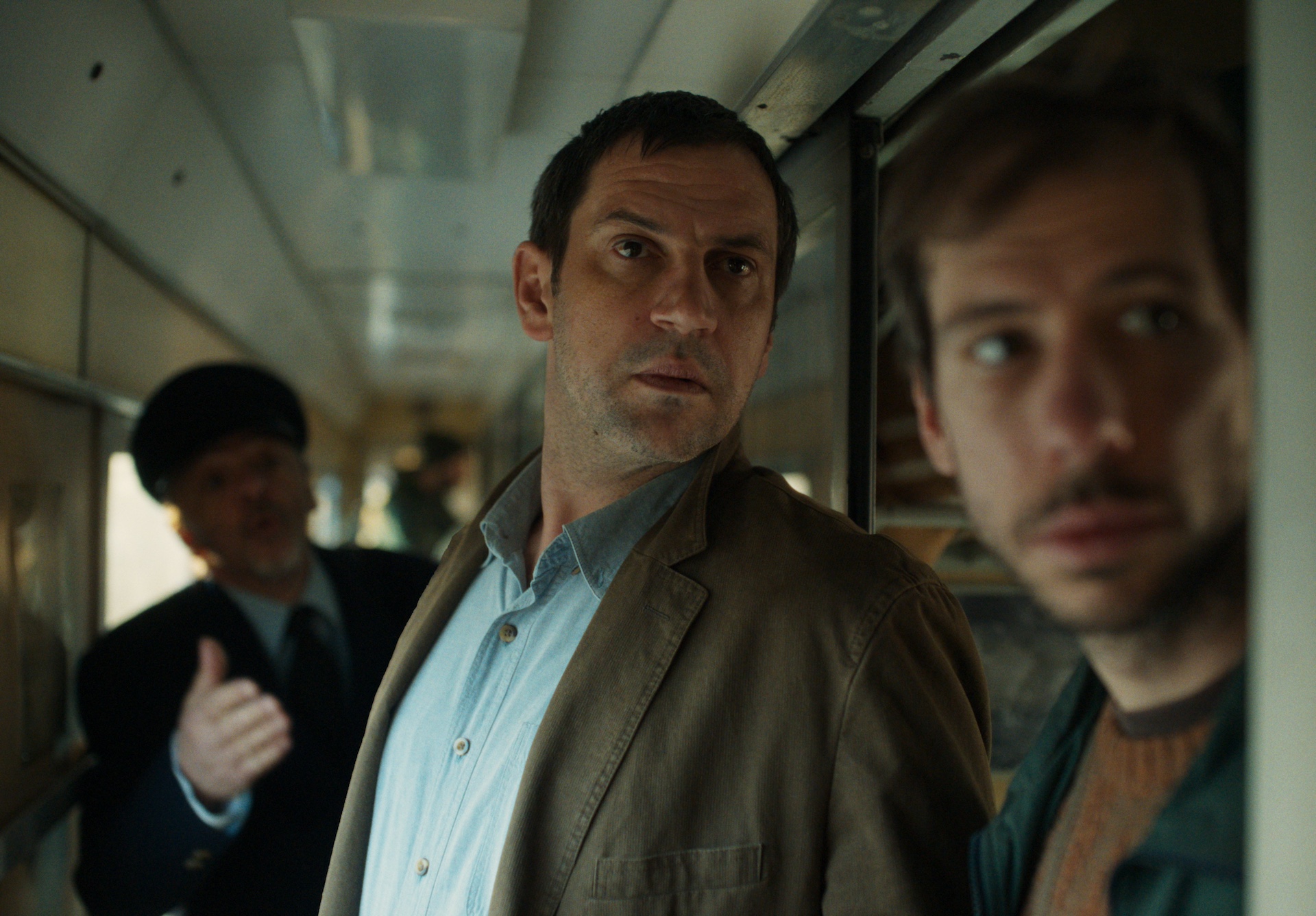 Three individuals stand closely together in a train compartment, with one gesturing as they converse amidst a crowded setting.
