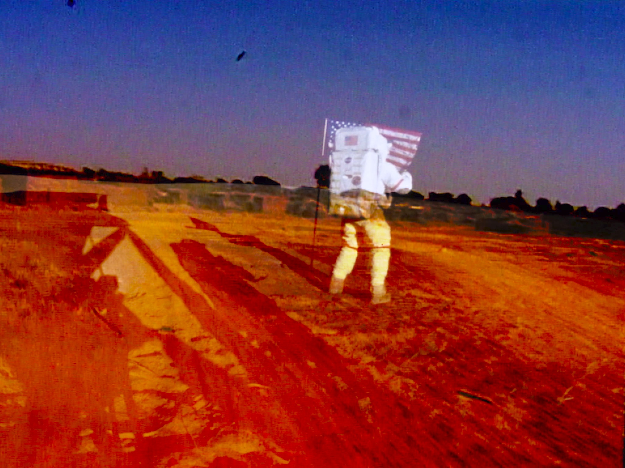 An astronaut in a spacesuit walks on a dusty surface, holding an American flag against a clear blue sky