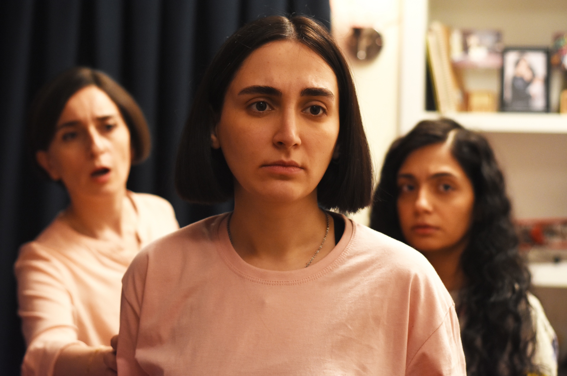 Two women stand behind a woman wearing a pink shirt in a softly lit room. Books and decor are visible in the background.